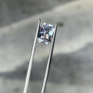 May include: A clear, square-shaped gemstone held by tweezers. The gemstone has a slight blue hue.