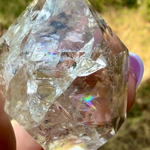 Puede incluir: Una punta de cristal de cuarzo transparente y facetada con fracturas internas e inclusiones que crean refracciones de luz arcoíris visibles en toda la piedra. El cristal tiene una parte superior puntiaguda y una superficie multifacetada.