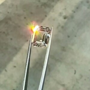 May include: A clear, square-shaped gemstone held by tweezers. The gemstone is reflecting light, creating a bright, white glow.