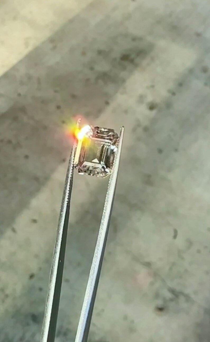 May include: A clear, square-shaped gemstone held by tweezers. The gemstone is reflecting light, creating a bright, white glow.