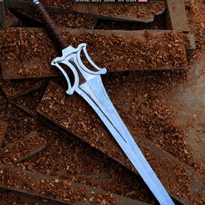 Puede incluir: Una espada de metal plateado con un mango envuelto en cuero marrón y una guarda de forma distintiva. La hoja presenta una cresta central y termina en punta. El texto POWER HEMAN SWORD es visible en la parte superior de la imagen.