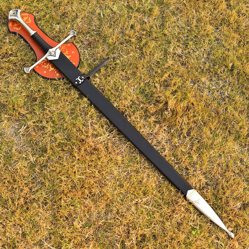 May include: A long sword with a black handle and blade, silver crossguard and pommel, and a leather sheath. The sword rests on a wooden plaque with an orange and brown design. The sword is displayed on a bed of grass.