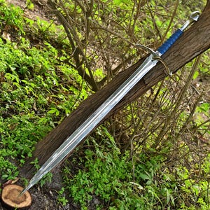 Puede incluir: Una espada larga y plateada con un mango envuelto en cuero azul y una guarda curvada. La hoja está muy pulida y refleja la vegetación circundante. La espada descansa sobre un tronco marrón, rodeada de follaje verde.
