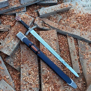 May include: A steel sword with a double-edged blade, a crossguard featuring intricate silver-toned metalwork, and a black grip. The pommel is circular with a red and silver emblem. It includes a black scabbard with silver-toned metal accents.