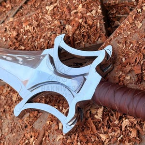May include: A close-up of a silver sword with a brown leather-wrapped handle. The sword has a distinctive guard design and is set against a background of wood shavings.