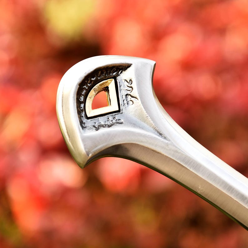 May include: Close-up of a silver-toned metal object with a gold-colored square inset. The object has a curved handle and a decorative design. The background is a blurred mix of red and green.