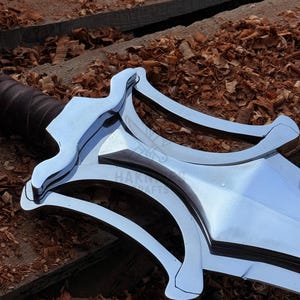 May include: Close-up of a polished metal sword with a distinctive curved guard. The blade is bright and reflective, with a dark brown leather-wrapped handle. The background features wood shavings and planks.
