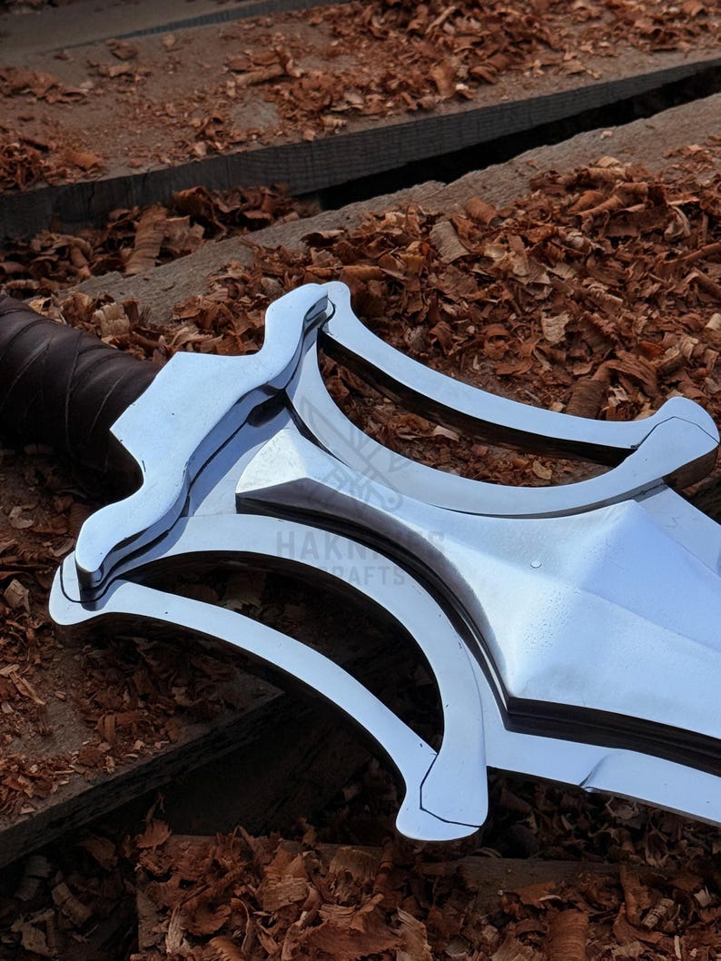 May include: Close-up of a polished metal sword with a distinctive curved guard. The blade is bright and reflective, with a dark brown leather-wrapped handle. The background features wood shavings and planks.