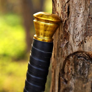 Puede incluir: Primer plano de una empu&ntilde;adura de espada con un pomo dorado y una empu&ntilde;adura envuelta en cuero negro. La empu&ntilde;adura se apoya contra un tronco de &aacute;rbol desgastado, con un fondo verde borroso.