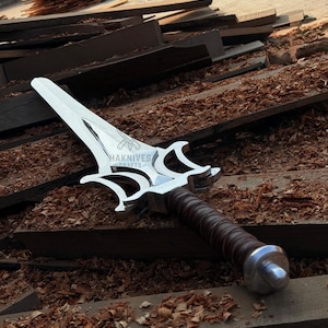 May include: A silver dagger with a decorative handle and a brown leather grip. The blade has a unique shape with a pointed tip and curved edges. The text "HAKNIVES CRAFTS" is visible on the blade.