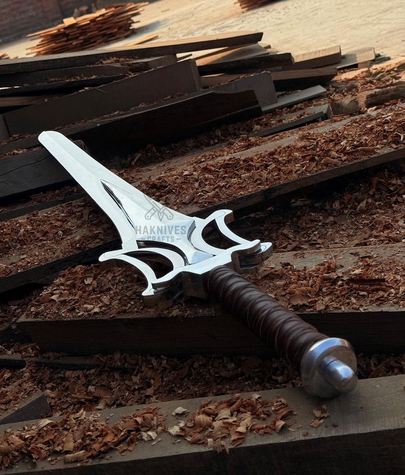 May include: A silver dagger with a decorative handle and a brown leather grip. The blade has a unique shape with a pointed tip and curved edges. The text "HAKNIVES CRAFTS" is visible on the blade.