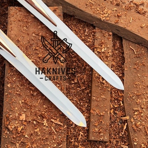May include: Two white swords with gold accents are displayed on a wooden surface covered in wood shavings. The swords have a sharp, pointed blade design. The text "HAKNIVES - CRAFTS" is visible.