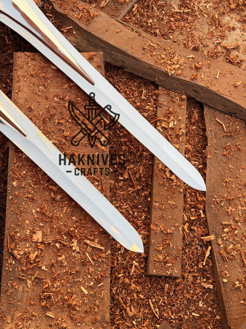 May include: Two white swords with gold accents are displayed on a wooden surface covered in wood shavings. The swords have a sharp, pointed blade design. The text "HAKNIVES - CRAFTS" is visible.