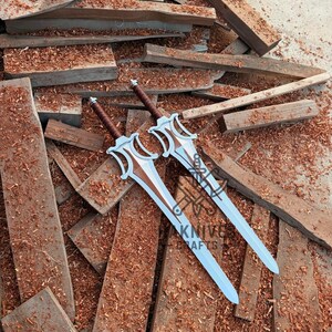 May include: Two decorative swords with silver blades and brown handles rest on a pile of wood. The swords have a distinctive design with a stylized hilt and a light blue blade. The text "KNIFE CRAFTS" is visible on the blade.