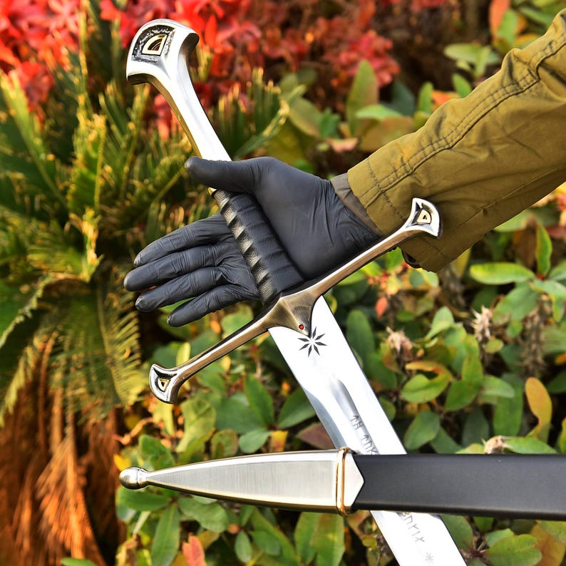 May include: A silver and gold sword with a black handle and a star design on the blade. The sword has a decorative hilt and is held against a backdrop of green foliage.