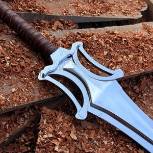 May include: A polished steel sword with a brown leather-bound handle. The blade reflects light. The sword rests on a wooden surface covered in wood shavings. A decorative weapon.