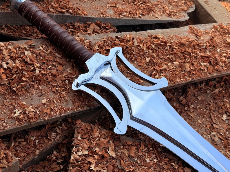 May include: A polished steel sword with a brown leather-bound handle. The blade reflects light. The sword rests on a wooden surface covered in wood shavings. A decorative weapon.