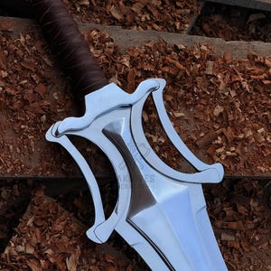 May include: A close-up of a silver sword with a brown leather-wrapped handle. The sword features a distinctive, stylized hilt with curved elements. The blade is polished and reflects light. The background is a wooden surface with wood shavings.