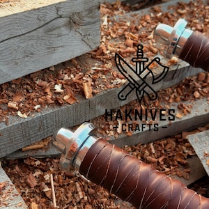May include: Close-up of two knife handles with brown leather wrapping and silver-colored metal accents. The handles are set against a background of wood shavings and wooden planks. The text "HAKNIVES CRAFTS" is visible.