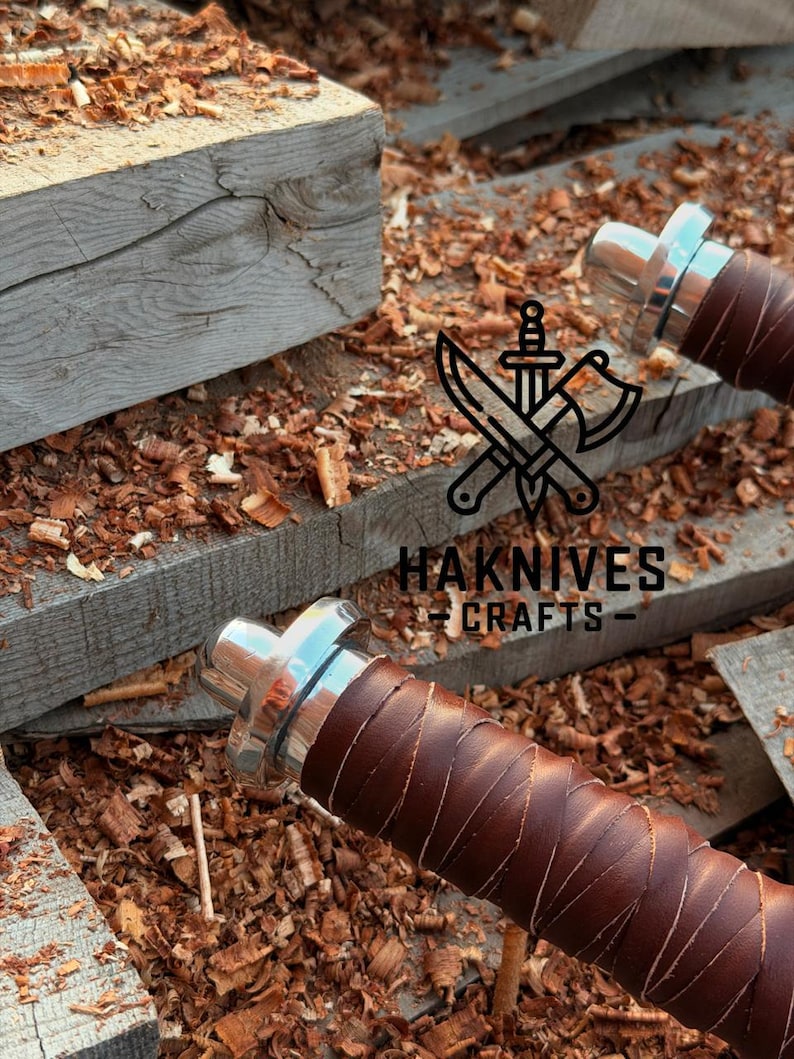 May include: Close-up of two knife handles with brown leather wrapping and silver-colored metal accents. The handles are set against a background of wood shavings and wooden planks. The text "HAKNIVES CRAFTS" is visible.