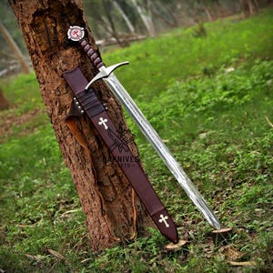 Puede incluir: Una espada medieval con una hoja plateada y un mango envuelto en cuero marrón. La espada está en una vaina de cuero marrón adornada con cruces blancas. La empuñadura presenta un emblema rojo y blanco. La espada está apoyada contra un tronco de árbol en una zona herbosa.
