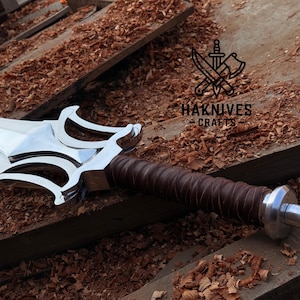 May include: A shiny steel sword with an ornate hilt and a brown leather-wrapped handle. The sword rests on a wooden surface with wood shavings. The text "HAKNIVES CRAFTS" is visible.