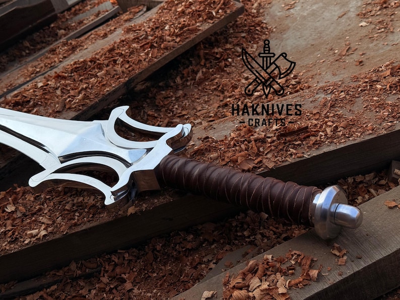May include: A shiny steel sword with an ornate hilt and a brown leather-wrapped handle. The sword rests on a wooden surface with wood shavings. The text "HAKNIVES CRAFTS" is visible.