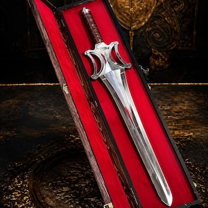Puede incluir: Una espada de metal plateado con una hoja ancha y puntiaguda y una guarda compleja y calada. El mango está envuelto en material marrón. La espada se exhibe dentro de un estuche de madera largo y rectangular forrado con tela de terciopelo rojo.