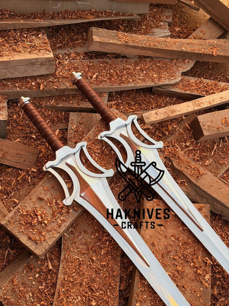May include: Two silver and brown swords with intricate designs, featuring brown leather-wrapped handles and a polished blade. The swords are displayed on a wooden surface, with the text "HAKNIVES - CRAFTS" visible.
