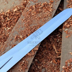 May include: A shiny, silver sword blade lies on aged wooden planks scattered with wood shavings. The blade is pointed and has a reflective surface. The logo "HAKNIV - CRA" is visible on the blade.