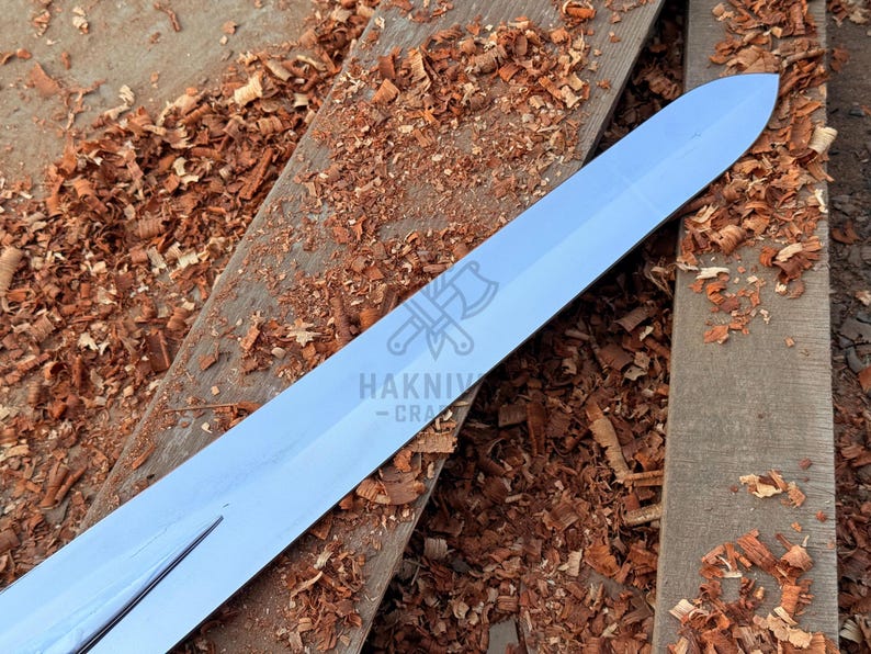 May include: A shiny, silver sword blade lies on aged wooden planks scattered with wood shavings. The blade is pointed and has a reflective surface. The logo "HAKNIV - CRA" is visible on the blade.