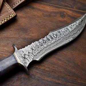 May include: A Damascus steel knife with a dark handle and a serrated edge. The blade features a unique pattern. A leather sheath with a button clasp is visible in the background. The knife rests on a wooden surface.