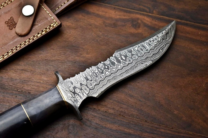 May include: A Damascus steel knife with a dark handle and a serrated edge. The blade features a unique pattern. A leather sheath with a button clasp is visible in the background. The knife rests on a wooden surface.