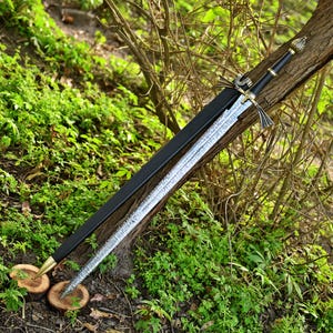 May include: A long sword with a patterned Damascus steel blade, a black handle, and a metallic crossguard. The sword is accompanied by a black scabbard featuring a gold-toned metal tip. The pommel is rounded with a textured, metallic finish.