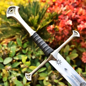 May include: A silver and bronze sword with a black handle. The blade has an etched starburst design and text. The hilt and pommel have a decorative design. The sword is set against a backdrop of green and red foliage.