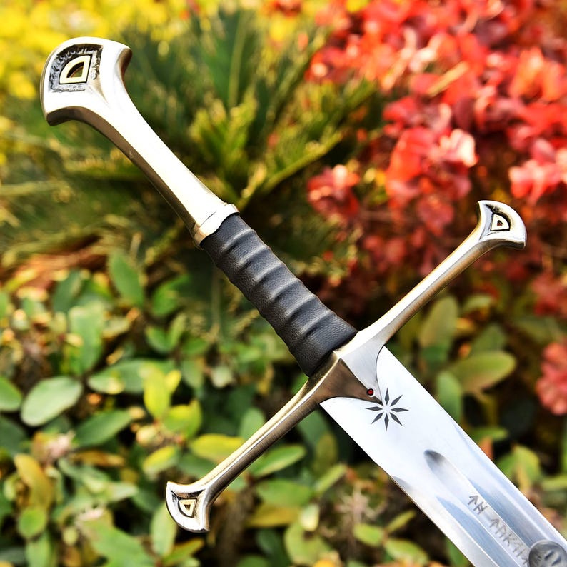 May include: A silver and bronze sword with a black handle. The blade has an etched starburst design and text. The hilt and pommel have a decorative design. The sword is set against a backdrop of green and red foliage.