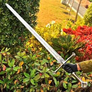 May include: A long, silver sword with a double-edged blade. The hilt and crossguard are silver with decorative accents. The blade has etched text and a star design near the hilt. The sword is held against a backdrop of green and red foliage.