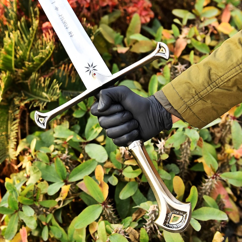 May include: A silver-colored sword with a decorative handle and crossguard. The blade has etched text and a star design. The handle is gold-colored with a decorative design. The sword is held in a gloved hand against a backdrop of green foliage.