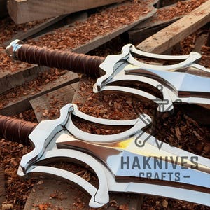 May include: Two polished metal swords with brown leather-wrapped handles. The blades are engraved with the text "HAKNIVES CRAFTS-". The swords are displayed on a wooden surface.