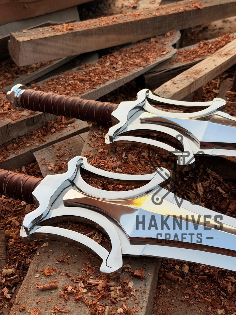 May include: Two polished metal swords with brown leather-wrapped handles. The blades are engraved with the text "HAKNIVES CRAFTS-". The swords are displayed on a wooden surface.