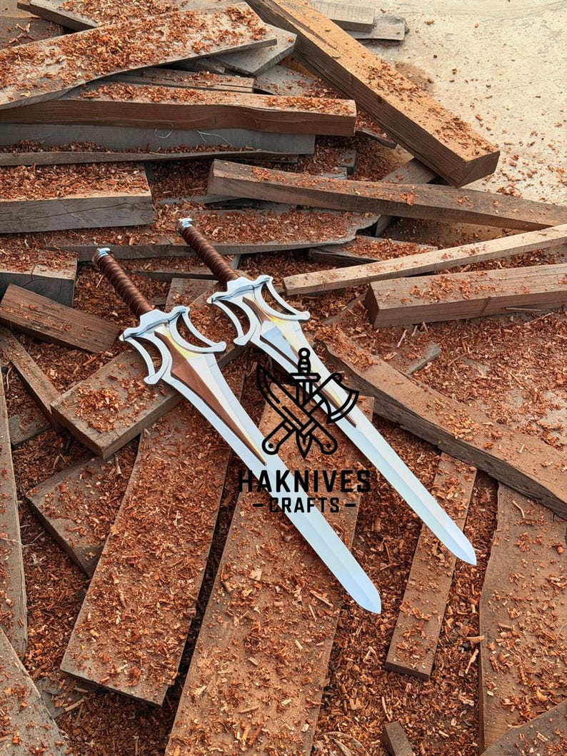 May include: Two silver and brown swords with ornate hilts rest on a pile of wooden planks and sawdust. The swords have a distinctive design with a curved guard and a brown leather-wrapped handle. The text "HAKNIVES CRAFTS" is visible.