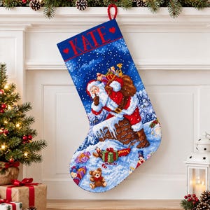 Op de afbeelding: Een blauwe kerstsok met een kruissteekontwerp van de Kerstman die uit een besneeuwde schoorsteen komt met een zak vol cadeaus. De naam KATE is in het rood bovenaan geborduurd, geflankeerd door twee kleine rode hartjes. Onderaan liggen een teddybeer en cadeautjes in de sneeuw.