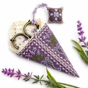 May include: A cone-shaped purple fabric scissor case featuring intricate cross-stitch floral patterns, trimmed with cream lace, and holding antique-style brass scissors. A small, matching square cross-stitched pincushion is attached by a braided cord.