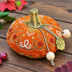 May include: A round, orange fabric pumpkin featuring intricate white, green, and red cross-stitch embroidery. It has a textured, light green ceramic stem, sage green cord accents, two smooth wooden beads, and a small green ceramic leaf charm.
