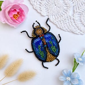 May include: A beaded beetle brooch featuring iridescent blue, green, and gold glass beads. The insect body is outlined in black seed beads with six black legs extending from the sides. A central vertical gold beaded stripe runs down the back.