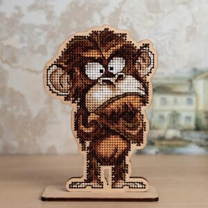 Può includere: Statuetta di scimmia in legno, ricamata a punto croce, con espressione accigliata. La scimmia è marrone e beige, con occhi bianchi e un motivo a punto dettagliato. È montata su una base di legno, ideale per l'esposizione.