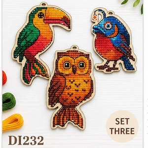 Könnte beinhalten: Set aus drei Vogelornamenten aus Holz mit Kreuzstichdetails. Enthält einen Tukan, einen blau-roten Vogel und eine Eule. Der Tukan hat einen großen orangen Schnabel, grüne Flügel und eine rote Brust. Die Eule ist braun und orange. Text "DI232" und "SET THREE".