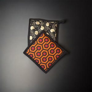 Peut inclure: Deux maniques carrées. L'une présente un fond noir avec un motif de crânes blancs et d'éléments botaniques. L'autre a un motif géométrique orange et rouge. Les deux ont des bordures noires et des boucles de suspension.