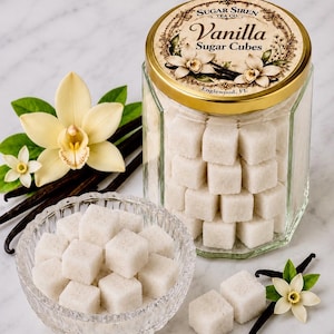 May include: A hexagonal glass jar filled with white vanilla sugar cubes, featuring a gold lid labeled Sugar Siren Tea Co. Vanilla Sugar Cubes Englewood, FL. A clear glass bowl contains additional sugar cubes, accompanied by vanilla flowers and dark pods.