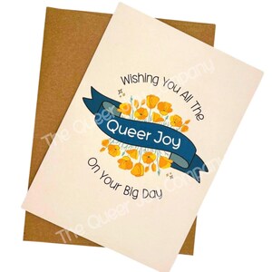 May include: A greeting card with a cream-colored background and a brown envelope. The card features a blue banner with the words "Queer Joy" and the text "Wishing You All The On Your Big Day". Yellow flowers surround the banner.